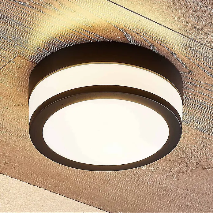 Lindby Deckenleuchte Flavi - schwarz, E27 - Runde Deckenleuchte mit dunklem Rahmen, weißem Diffusor und leuchtendem Seitenring, auf einer Holzdecke strahlend.