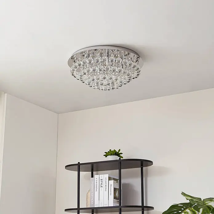 Lindby Deckenleuchte Antonino - chrom / silber, klar / transparent, G9 - Ein flächenbündiger Kristall-Kronleuchter mit silberner Basis beleuchtet einen modernen Raum, über einem dunklen Regal.