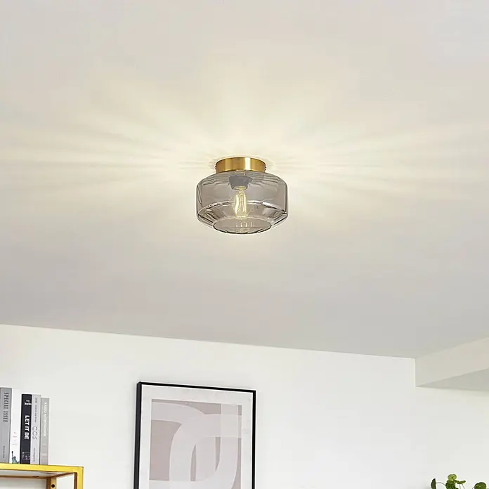 Lindby Deckenleuchte Hildur - L x B x H: 22 x 22 x 15,3 cm, alu / grau / zink, E27 - Eine messingfarbene und gerippte graue Glas-Deckenleuchte, leuchtend mit Lichtmustern an einer weißen Decke.