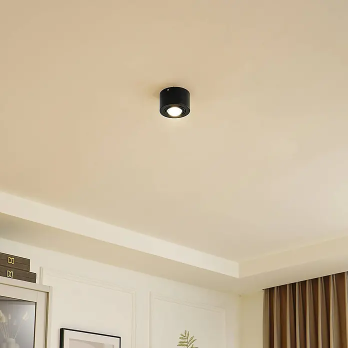 Lindby Deckenlampe in schwarz matt, Deckenleuchte -  - Ein schwarzer zylindrischer Deckenstrahler, an einer hellen Decke montiert, strahlt warmweißes Licht aus.