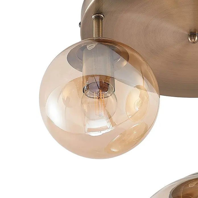 Lindby Deckenleuchte Teeja - bronze / altmessing, creme / amber, E27 - Nahaufnahme einer messingfarbenen Deckenleuchte mit kugelförmigem, amberfarbenem Glasschirm und sichtbarer Glühbirne.