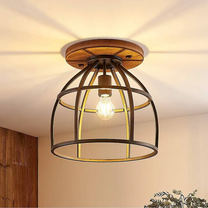 Lindby Deckenleuchte Rutger - Holz dunkel, schwarz, E27 - Industrielle Deckenleuchte mit dunklem Metallkäfig, goldenen Akzenten und Holzplatte, wirft warmes Licht und Schatten.