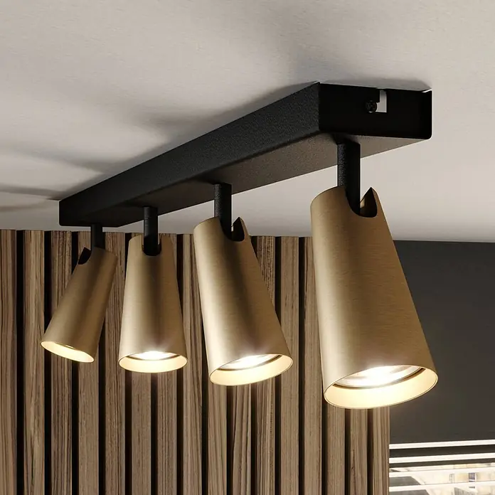 Lucande Deckenstrahler Angelina - schwarz, messing / gold, GU10 - Vierflammige Deckenleuchte mit schwarzer Metallschiene und konischen Holzschirmen, die warmes Licht auf eine Holzwand wirft.
