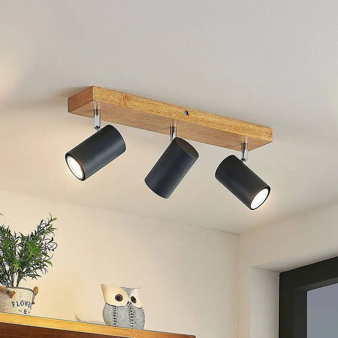 Lindby Deckenstrahler Maniva - Holz hell, schwarz, GU10 - Drei schwarze zylindrische Strahler mit Chromhalterungen auf einem rechteckigen Holzsockel sind an der Decke befestigt und beleuchten den Raum.