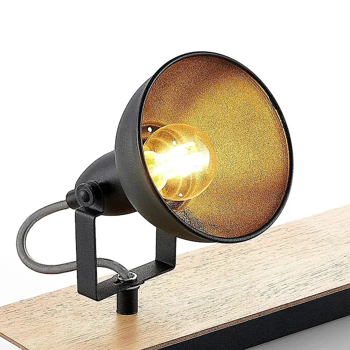 Lindby Deckenstrahler Aylis - Holz dunkel, schwarz, E14 - Schwarze Metall-Spotlampe mit goldener Innenseite und leuchtender Glühbirne, montiert auf einem hellen Holzsockel mit dunklem Rand.