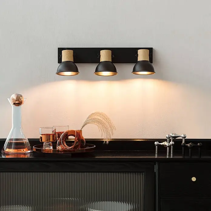 Lindby Deckenstrahler Bosko - schwarz, E14 - Drei-Flammen-Wandleuchte mit schwarzen Metallschirmen und Holzdetails, die warmes Licht auf eine dunkle Theke wirft.
