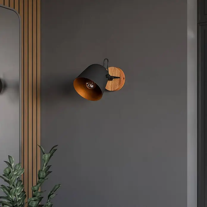 Lindby Deckenstrahler Adalin - L x B x H: 12 x 12 x 15,7 cm, Holz dunkel, schwarz, E14 - Schwarze Metall-Wandleuchte mit sichtbarer Glühfadenbirne, goldenem Innenraum und rundem Holzsockel, an dunkler Wand.