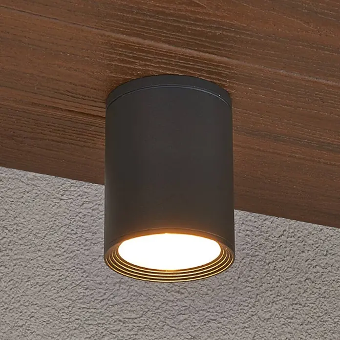 Lucande Deckenstrahler Minna - schwarz, E27 - Eine schwarze zylindrische Aufbauleuchte mit warmem Licht, montiert an einem Holzbalken.