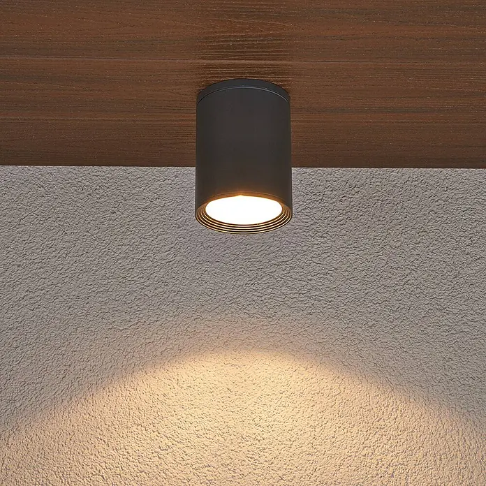 Lucande Deckenstrahler Minna - schwarz, E27 - Dunkle zylindrische Deckenleuchte, an Holzbalken montiert, wirft warmes Licht auf eine strukturierte Wand.