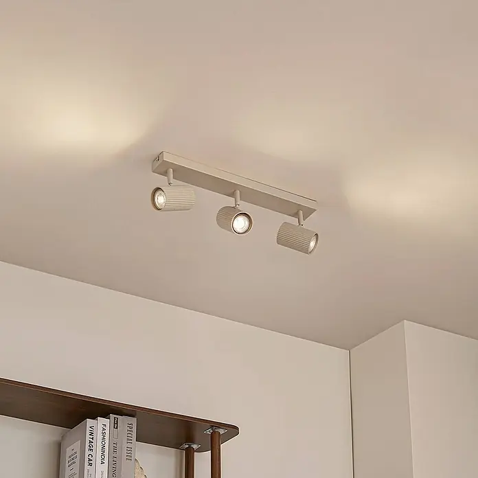 Lindby Deckenstrahler Kardis - L x B x H: 45 x 6 x 16 cm, creme / amber, creme / amber, GU10 - Beige Deckenleuchte mit drei gerippten Strahlern, die einen Raum über einem Bücherregal beleuchtet.