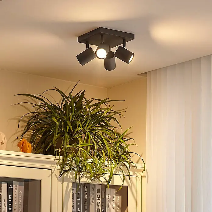 Lindby Deckenstrahler Jorell - schwarz, schwarz, GU10 - Schwarzer Deckenstrahler mit vier verstellbaren Köpfen über einem Bücherregal mit Pflanze, der warmes Licht spendet.