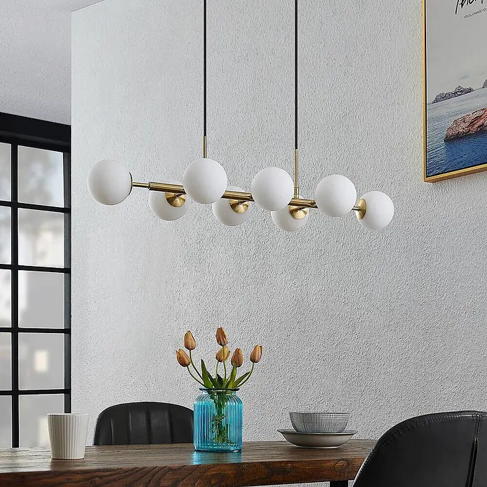 Lindby Pendelleuchte Kiamo - L x B x H: 82,5 x 32,5 x 10 cm, weiß / opal, messing matt, opalweiß, G9 - Eine moderne Pendelleuchte mit goldfarbenem Gestell und weißen Glaskugeln hängt über einem Esstisch.