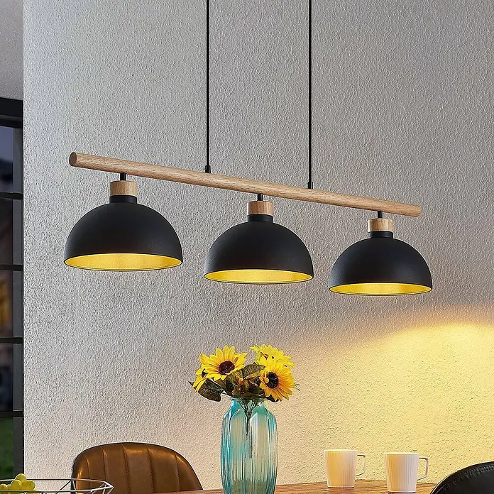 Lindby Pendelleuchte Tirzana - L x B x H: 100 x 25 x 12,5 cm, schwarz, Schwarz, gold, holz hell, E27 - Lineare Pendelleuchte mit drei schwarzen Metallschirmen, goldfarbenen Innenseiten und einer Naturholzstange, über einem Esstisch.