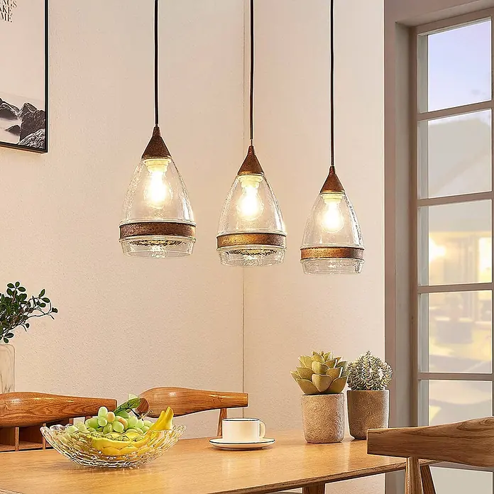 Lindby Pendelleuchte Millina - L x B x H: 70,4 x 15,6 x 25,8 cm, klar / transparent, rostbraun, klar, E27 - Drei Pendelleuchten mit klaren, strukturierten Glasschirmen und Bronzeakzenten beleuchten einen Esstisch.