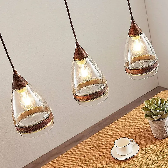 Lindby Pendelleuchte Millina - L x B x H: 70,4 x 15,6 x 25,8 cm, klar / transparent, rostbraun, klar, E27 - Drei Pendelleuchten mit strukturierten Glasschirmen und kupferfarbenen Fassungen über einem Holztisch.