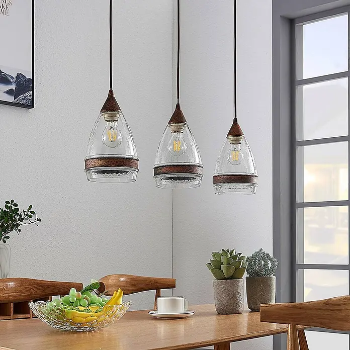 Lindby Pendelleuchte Millina - L x B x H: 70,4 x 15,6 x 25,8 cm, klar / transparent, rostbraun, klar, E27 - Drei Pendelleuchten mit klaren, strukturierten Glasschirmen und rustikalen Bronzeakzenten über einem Esstisch.