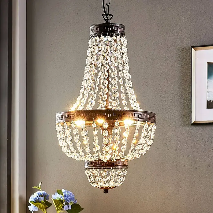 Lindby Pendelleuchte Jorve - L x B x H: 35 x 35 x 150 cm, klar / transparent, Schwarz gold gebürstet, klar, G9 - Verzierter bronzefarbener Kronleuchter mit kaskadierenden Kristallperlen, hell beleuchtet, in einem Raum hängend.