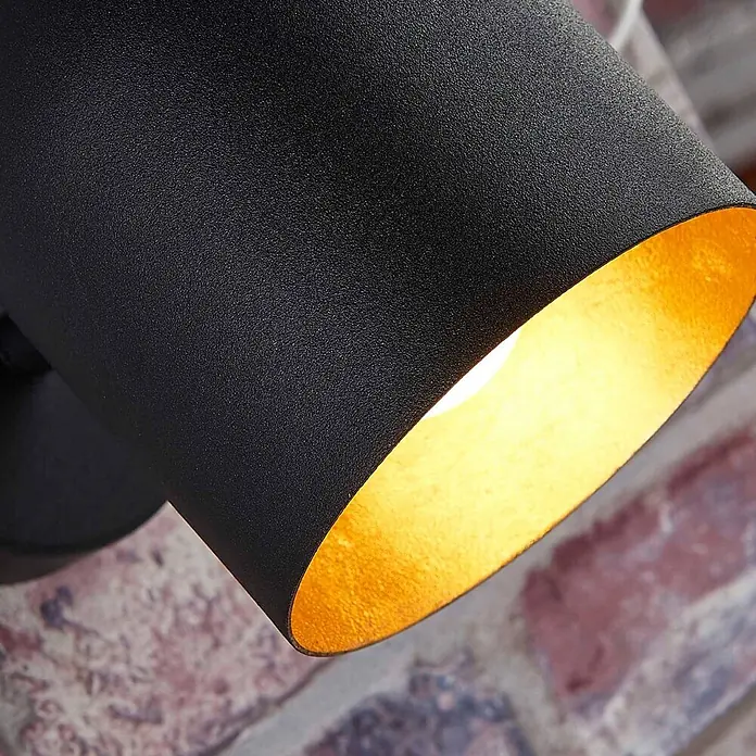 Lindby Deckenstrahler Morik - L x B x H: 19 x 10 x 12 cm, schwarz, schwarz, E14 - Nahaufnahme einer schwarzen strukturierten Lampe mit goldener Innenseite, beleuchtet, an einer Ziegelwand montiert.