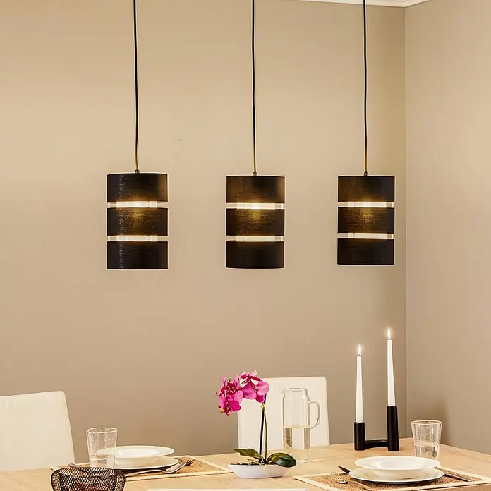 Lindby Pendelleuchte Tsomo - L x B x H: 950 x 18 x 28 cm, schwarz, Schwarz, gold, E27 - Drei schwarze zylindrische Pendelleuchten mit beleuchteten Bändern hängen über einem Esstisch.