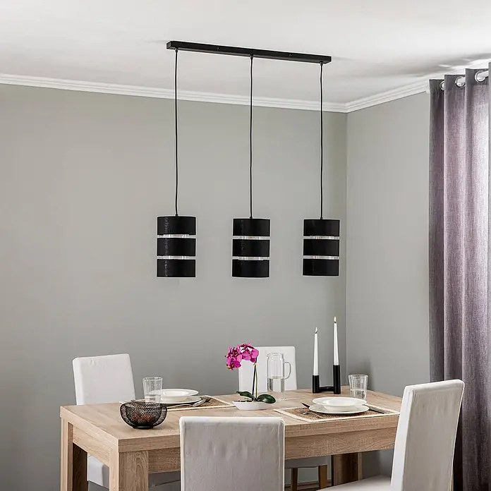 Lindby Pendelleuchte Tsomo - L x B x H: 950 x 18 x 28 cm, schwarz, Schwarz, gold, E27 - Dreiflammige Pendelleuchte mit schwarzen zylindrischen Schirmen und silbernen Akzenten, an einer schwarzen Stange über einem Esstisch hängend.