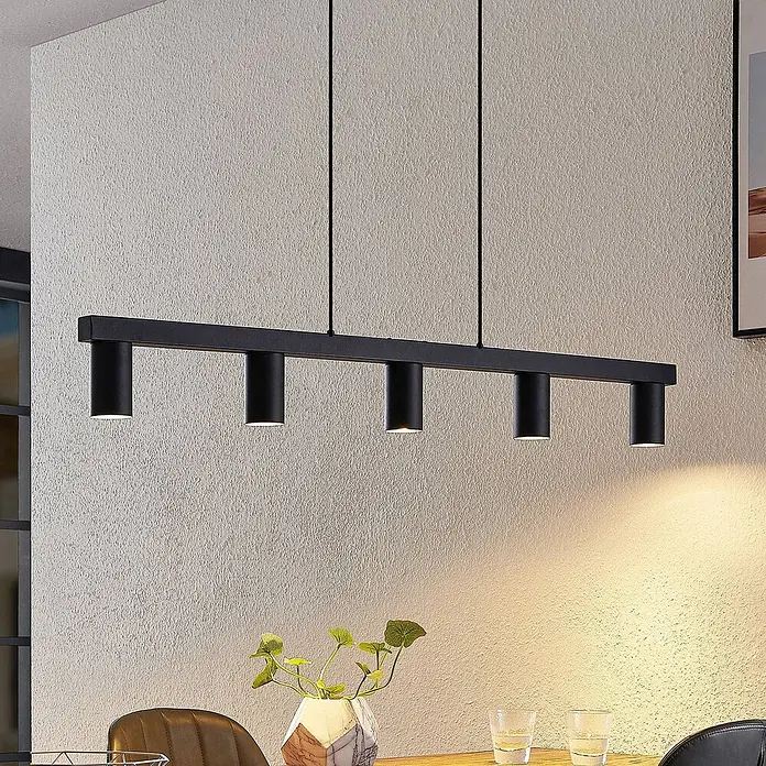 Lindby Pendelleuchte Baliko - schwarz, Schwarz, GU10 - Eine schwarze lineare Metall-Pendelleuchte mit fünf zylindrischen Strahlern hängt über einem Holztisch und beleuchtet ihn warm.