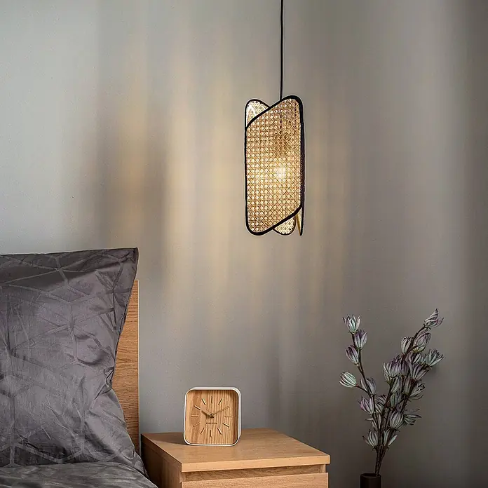 Lucande Pendelleuchte Bassiola - L x B x H: 19 x 19 x 37 cm, Holz hell, Schwarz, Holz hell, E27 - Eine beleuchtete Pendelleuchte mit schwarzem Rahmen und Rattanschirm hängt über einem Nachttisch und spendet sanftes Licht.