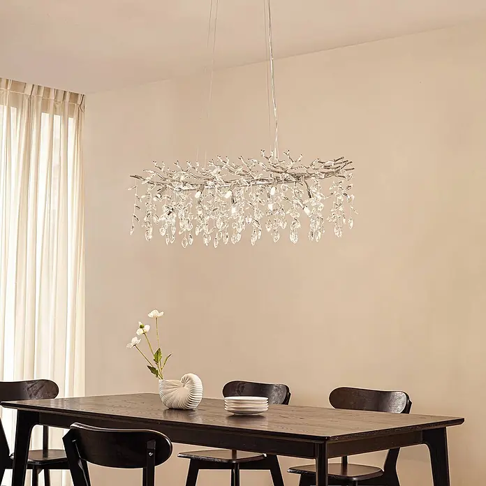 Lucande Pendelleuchte Bellora - L x B x H: 101 x 45 x 32 cm, klar / transparent, chrom, transparent, G9 - Rechteckiger Kronleuchter mit silberfarbenen Ästen und klaren Kristalltropfen, über einem dunklen Holzesstisch in einem modernen Raum.