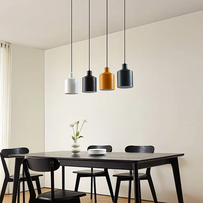 Lindby Pendelleuchte Tirane - L x B x H: 94 x 16 x 13 cm, Multicolour, Schwarz, weiß, Grün, gelb, E27 - Vier industrielle Pendelleuchten aus Metall in Weiß, Schwarz, Orange und Dunkelgrau hängen über einem schwarzen Esstisch aus Holz.
