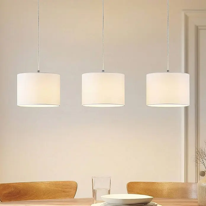 Lindby Pendelleuchte Imarin - L x B x H: 100 x 25 x 18 cm, weiß / opal, Holz hell, weiß, E27 - Drei moderne Pendelleuchten mit weißen zylindrischen Stoffschirmen hängen über einem hellen Holztisch mit Tellern und einem Glas.