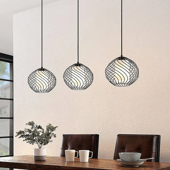 Lindby Pendelleuchte Cedrice - L x B x H: 108 x 25 x 21 cm, schwarz, Schwarz, weiß, E27 - Drei Pendelleuchten mit schwarzen Metallspiralgeflechten und weißen Innenkugeln hängen über einem Holztisch.