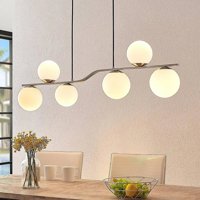 Lindby Pendelleuchte Avalyn - L x B x H: 110 x 16 x 32 cm, weiß, nickel satiniert, E14 - Moderne Pendelleuchte mit sechs kugelförmigen weißen Glasschirmen an einem gewellten Goldstab über einem Holztisch.