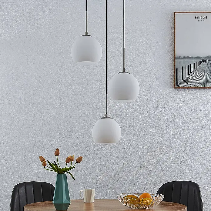 Lindby Pendelleuchte Jurian - L x B x H: 32 x 32 x 17,5 cm, weiß / opal, opal, nickel, E27 - Drei weiße kugelförmige Pendelleuchten mit mattem Glas und dunklen Metallstäben hängen über einem Esstisch mit Stühlen, Vase und Obst.