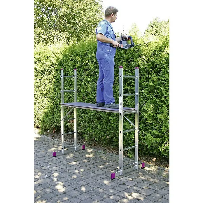 Krause Corda Leitergerüst - Arbeitshöhe: 3,75 m, 12 Stk. - Eine Person auf einem Gerüst schneidet mit einer Heckenschere eine Hecke, wobei das silberne Gerüst mit lila Akzenten hervorsticht.