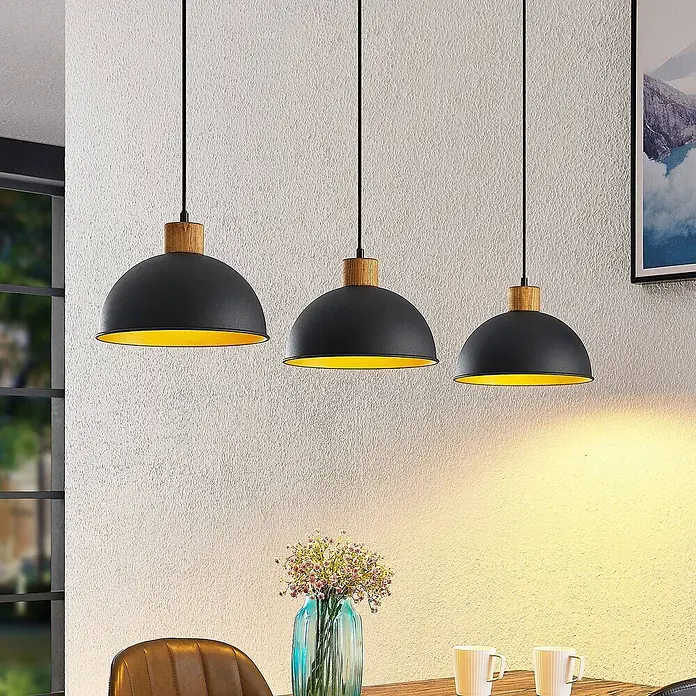 Lindby Pendelleuchte Flurin - schwarz, sandSchwarz, gold, holz, E27 - Drei schwarze Hängeleuchten mit Holzdetails und goldener Innenseite hängen über einem Esstisch.