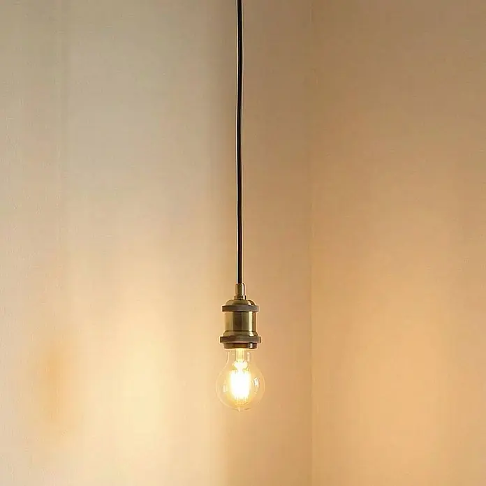 Lindby Pendelleuchte Hattie - messing / gold, messing, E27 - Messing-Pendelleuchte mit sichtbarer Glühbirne, hängt und wirft warmes Licht auf eine Wandecke.