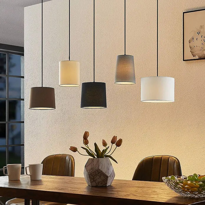 Lindby Pendelleuchte Amilia - Multicolour, Holz natur, Schwarz, grau, beige, braun, weiß, E27 - Fünf Pendelleuchten aus Stoff in verschiedenen Formen und Farben hängen über einem Holztisch.