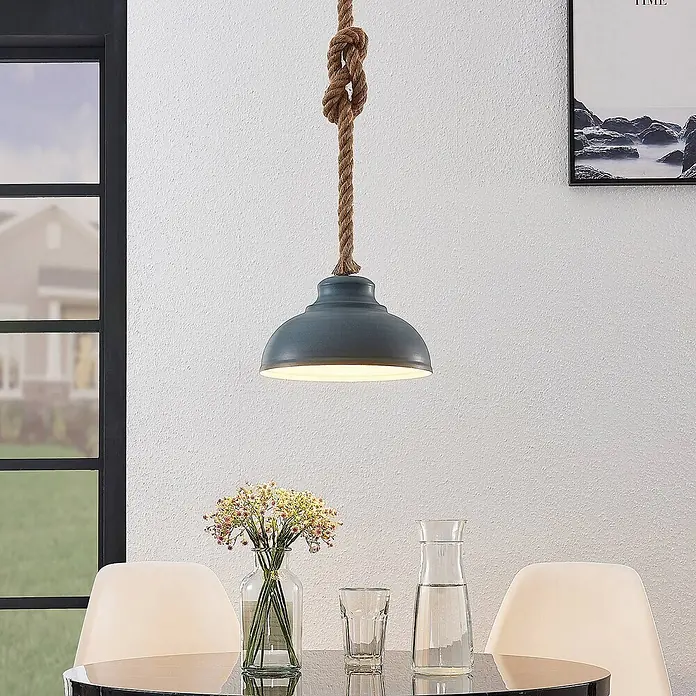 Lindby Pendelleuchte Chaby - alu / grau / zink, beton, E27 - Eine blau-graue Metall-Pendelleuchte mit geknoteter Seilaufhängung hängt über einem schwarzen Tisch mit Blumen und Glaswaren.