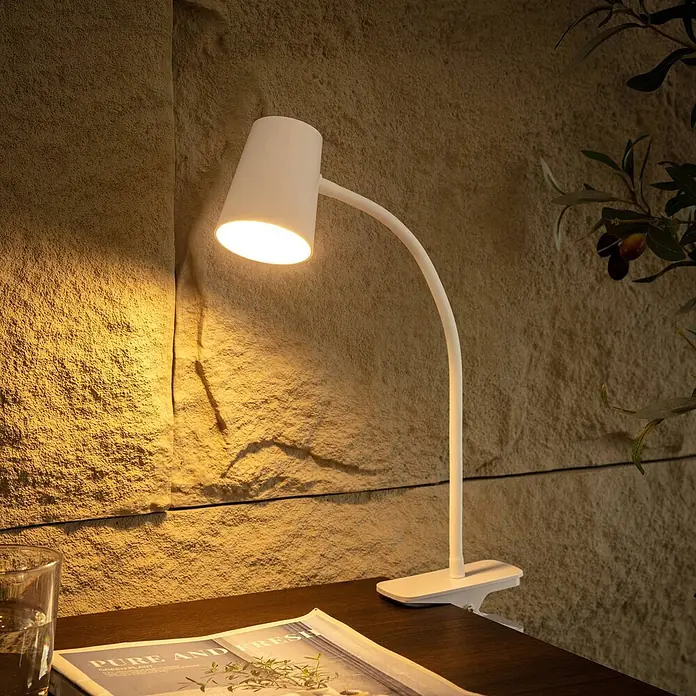 Lindby Klemmleuchte Ailina - Farbe Gehäuse: weiß / opal - Weiße Schwanenhals-Klemmleuchte beleuchtet eine strukturierte Wand auf einem dunklen Schreibtisch.