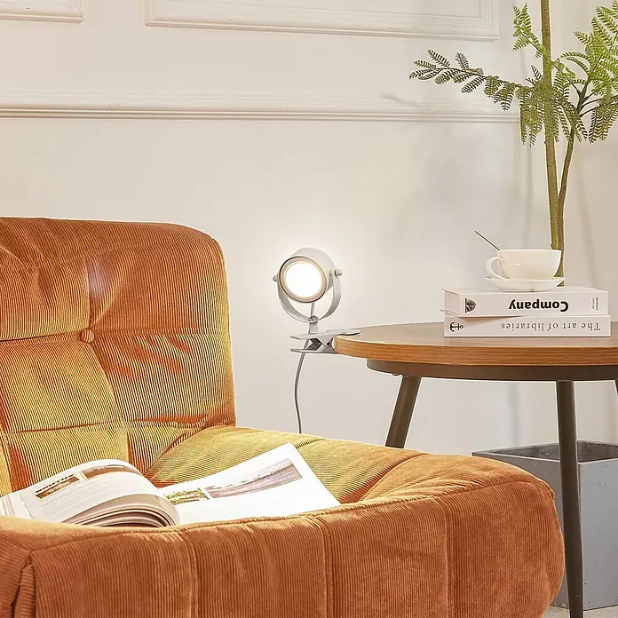 Lindby Klemmleuchte Jyla - Farbe Gehäuse: weiß / opal - Graue Klemmlampe beleuchtet ein offenes Buch auf einem orangefarbenen Kord-Sessel, an einem Holztisch befestigt.