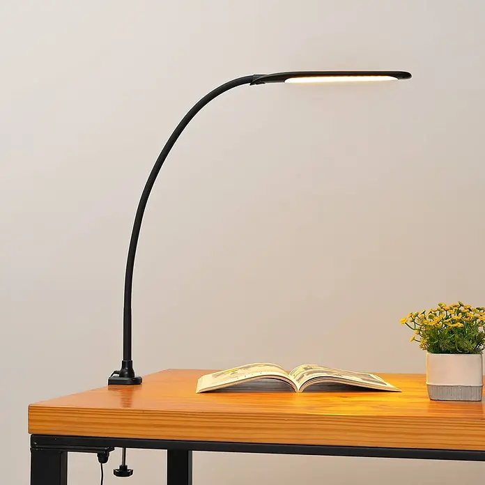 Lindby Klemmleuchte Nilay - Farbe Gehäuse: schwarz - Schwarze LED-Schreibtischlampe mit gebogenem Arm, an Holzschreibtisch geklemmt, beleuchtet ein offenes Buch.