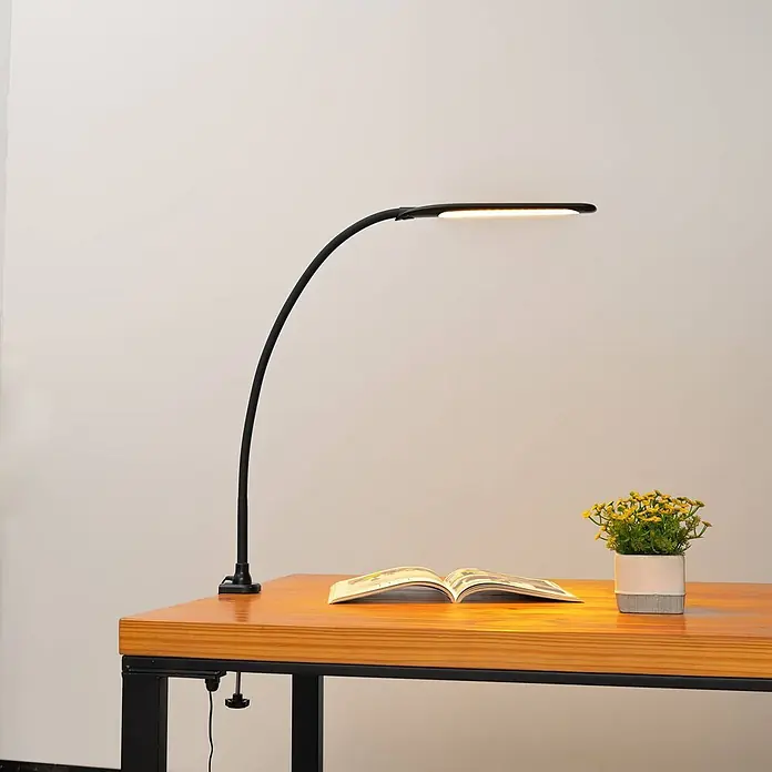 Lindby Klemmleuchte Nilay - Farbe Gehäuse: schwarz - Schwarze Schreibtischlampe mit gebogenem Arm und LED-Lichtleiste, an einem Holztisch befestigt, beleuchtet einen Leseplatz.
