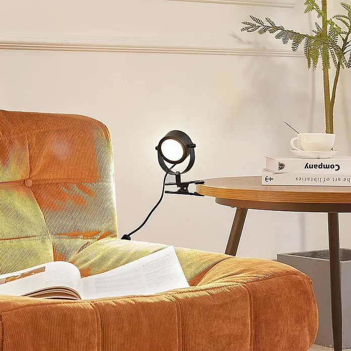 Lindby Klemmleuchte Jyla - Farbe Gehäuse: schwarz - Schwarze Klemmleuchte aus Metall mit hellem Licht, an einem Holznebentisch neben einem orangefarbenen Sessel mit Buch.