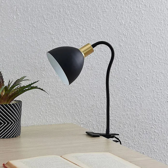 Lindby Klemmleuchte Genora - L x B x H: 31,5 x 12 x 21 cm, Farbe Gehäuse: schwarz - Schwarze Klemmleuchte mit Goldakzent, auf hellem Holztisch neben Pflanze und offenem Buch.