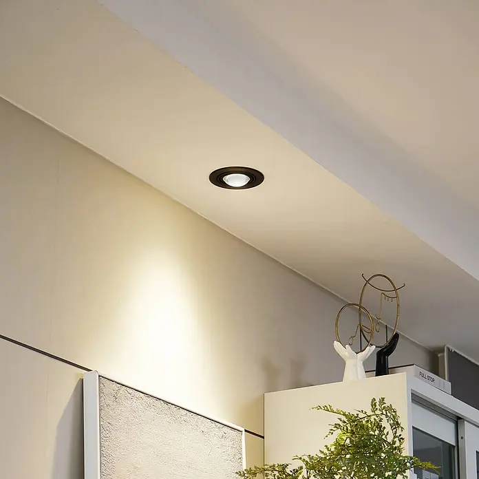 Lindby Einbauleuchte Jyla - Ø x H: 13 x 5,5 cm, schwarz - Dunkel umrandeter Einbaustrahler in einer Decke, der eine Wand beleuchtet.