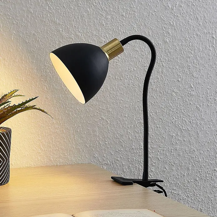 Lindby Klemmleuchte Genora - L x B x H: 31,5 x 12 x 21 cm, Farbe Gehäuse: schwarz - Schwarze Klemm-Schreibtischlampe mit goldenem Akzent und flexiblem Arm, die einen Holztisch neben einer Pflanze beleuchtet.