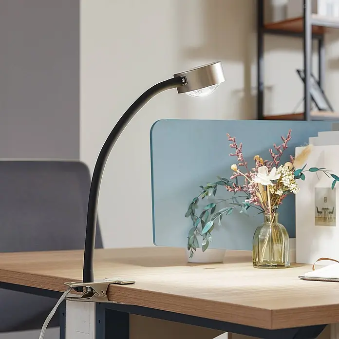 Lindby Klemmleuchte Jyla - L x B x H: 30 x 8 x 25 cm, Farbe Gehäuse: alu / grau / zink - Schwarze flexible Klemm-Schreibtischlampe mit Schwanenhals und gebürstetem Metallkopf, die einen Holzschreibtisch beleuchtet.