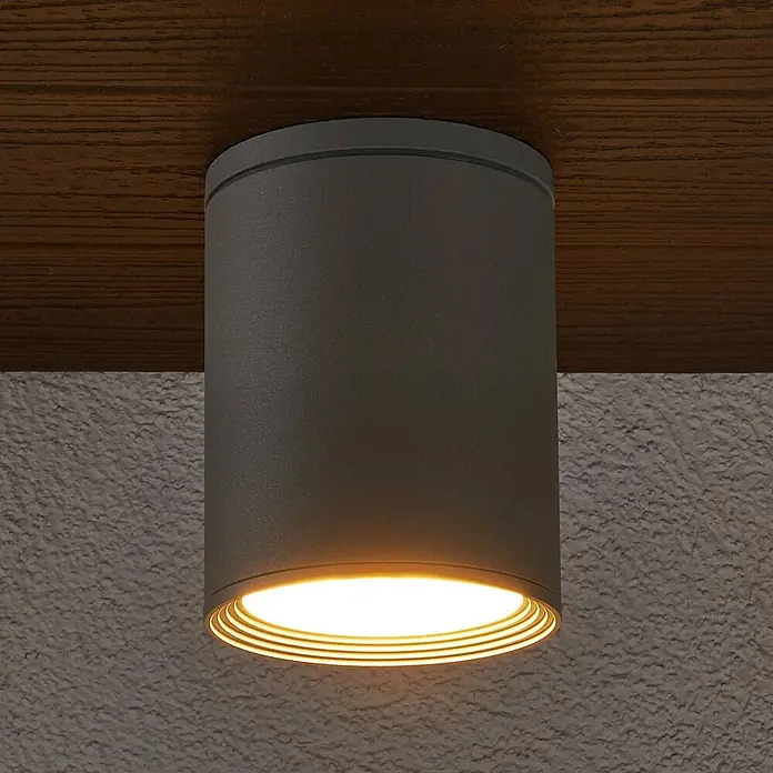 Lucande Außenwand- & Deckenleuchte Minna - alu / grau / zink, alu / grau / zink, IP54 - Dunkelgraue zylindrische Deckenleuchte mit warmer Beleuchtung, an einer Holzdecke über einer strukturierten Wand montiert.