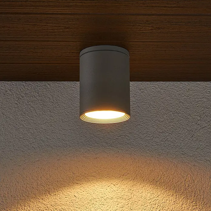 Lucande Außenwand- & Deckenleuchte Minna - alu / grau / zink, alu / grau / zink, IP54 - Dunkelgraue zylindrische Deckenleuchte an Holzdecke, spendet warmes Licht auf strukturierte Wand.