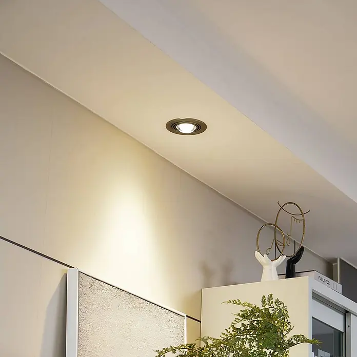 Lindby Einbauleuchte Jyla - alu / grau / zink - Ein Einbaustrahler mit dunkler Metallblende ist in einer Decke installiert und wirft warmes Licht auf die darunterliegende Wand.