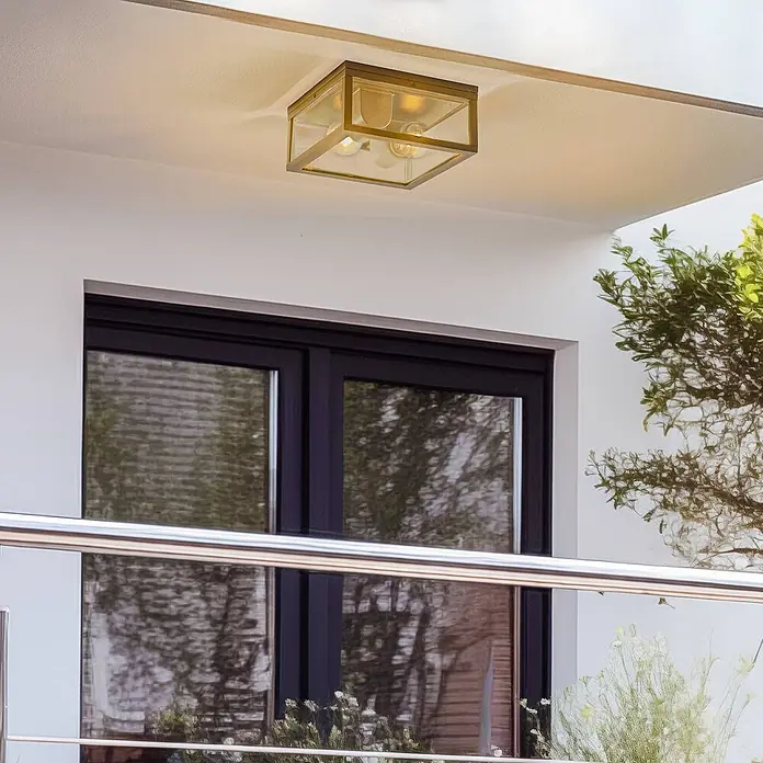 Lindby Außenwand- & Deckenleuchte Kremena - schwarz, klar / transparent, IP44 - Quadratische, messingfarbene Deckenleuchte mit klaren Glaspaneelen, leuchtet einen Bereich über einem dunklen Fenster aus.